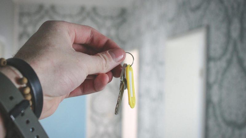 A hand holding a key against a blurred background of patterned wallpaper indoors.